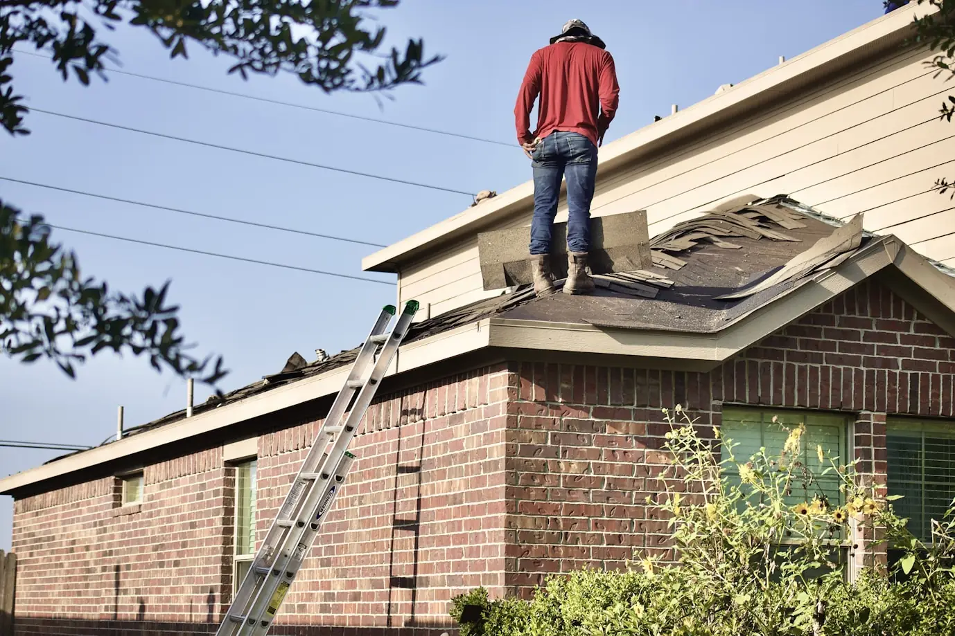 Worker tearing off old shingles above a brick and siding home with a ladder against the eave